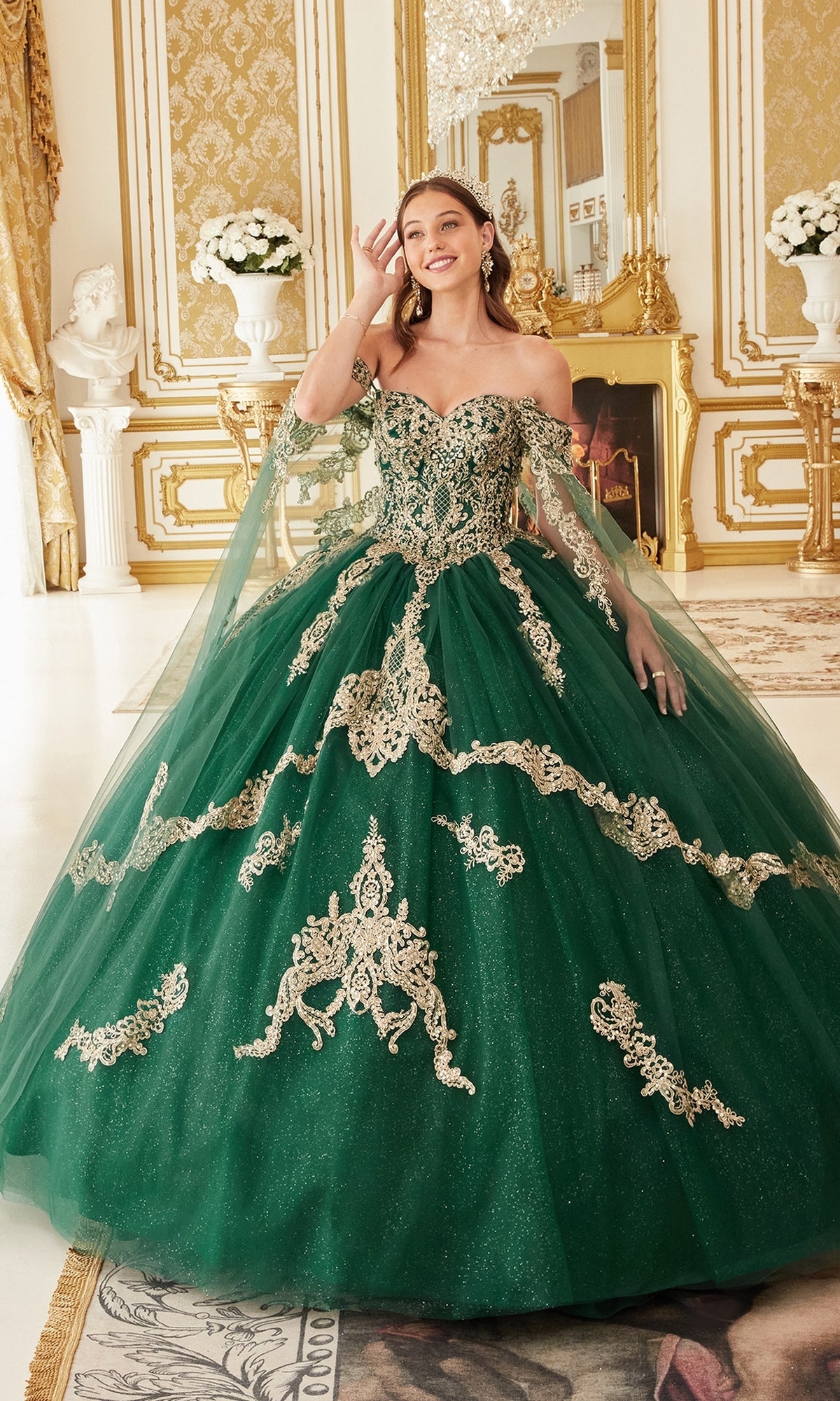 A woman stands in an elegant room wearing the Ladivine 15711 Quinceañera Dress, featuring emerald green fabric, gold embroidery, off-the-shoulder sleeves, and a full glitter tulle skirt.